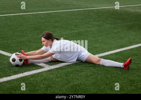 Ein junger Fußballspieler dehnt sich auf einem grünen Feld aus. Sie ist in einer gespaltenen Position und greift mit beiden Händen nach einem Fußball. Vorbereitung auf ein Spiel. Stockfoto