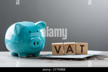 Die MwSt. ist auf einem Holzblock neben einem Münzhaufen, einer blauen Schweinebank und einem Taschenrechner auf einem Holztisch vor grauem Hintergrund geschrieben. Stockfoto