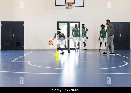 Dribbelt Basketball um Kegel, Spieler üben, während Teamkollegen und Coach zusehen Stockfoto