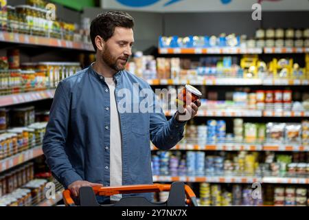 Mann im Lebensmittelgeschäft, der beim Einkaufen Konserven hält. Umgeben von verschiedenen Speisen. Einkaufskonzept, Kaufentscheidungen und Lebensmittelverfügbarkeit. Stockfoto