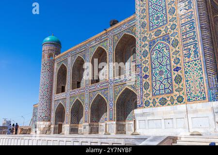 Samarkand, Usbekistan - 25. März 2024: Tilya-Kori Madrasah-Fassade an einem sonnigen Tag. Touristen laufen auf dem Registan-Platz Stockfoto