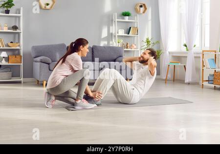 Glückliche, sportliche junge Frau, die ihrem Mann hilft, zu Hause sportliche Fitnesstraining zu machen. Stockfoto