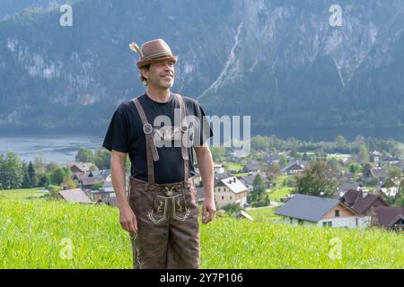 Ein älterer Bauer in Nationalkleidung, Ledershorts und einer Kappe mit Feder steht vor einem Hintergrund von Bergen und grünem Gras. Stockfoto