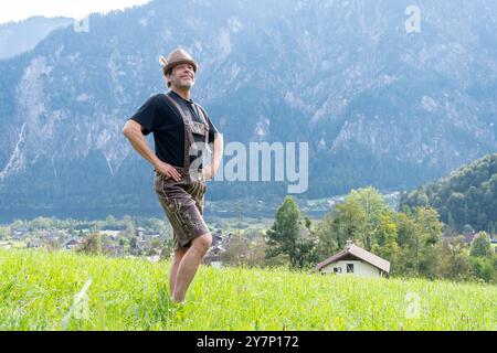 Ein älterer Bauer in österreichischer Nationalkleidung tanzt vor einer Kulisse aus Bergen und einer kleinen Stadt. Stockfoto