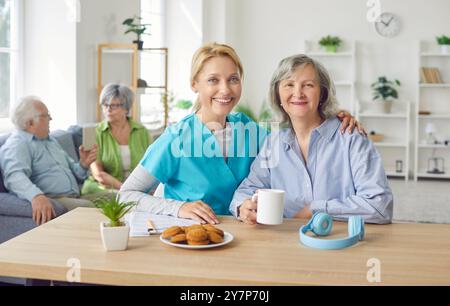 Eine Krankenschwester, die an einem Tisch sitzt, umarmt eine ältere Frau in einer Pension, Porträt Stockfoto