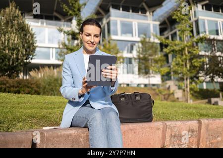 Glückliche Geschäftsfrau, die im Freien ein digitales Tablet nutzt und einen sonnigen Tag mit modernen Bürogebäuden im Hintergrund genießt Stockfoto