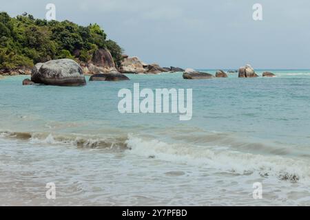 Eine ruhige Strandszene mit großen Felsbrocken im flachen Wasser, mit schaumigen Wellen, die an einem sonnigen Tag zum Ufer Rollen. Stockfoto