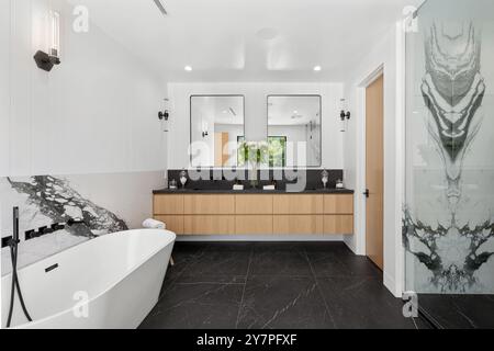 Modernes Bad mit freistehender Badewanne, zwei Waschbecken, großen Spiegeln und einer Glasdusche mit Marmorakzenten und schwarzen Bodenfliesen. Stockfoto