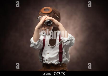 Kinderpilot mit Fliegermütze und Piratenfernglas Spielzeug, Retro-Porträt eines Jungen in Vintage-Kleidung vor altem braunen Hintergrund Stockfoto