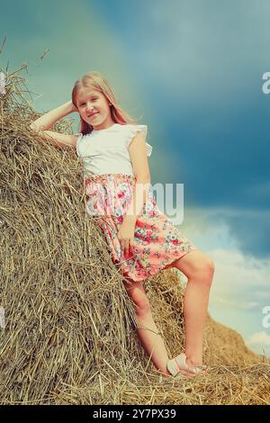 Ein blondes Mädchen mit langen lockeren Haaren in einem rosa Kleid kletterte auf große Strohballen Stockfoto