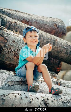 Ein kleiner Junge in Baseballmütze und Jeansshorts sitzt auf Baumstämmen und lacht fröhlich Stockfoto