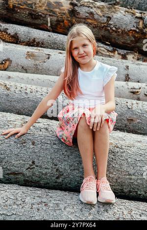 Ein blondes Mädchen mit langen, lockeren Haaren, lächelnd, sitzt in einem rosa Kleid auf Baumstämmen Stockfoto