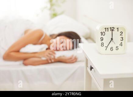 Junge, friedliche Frau, die zu Hause unter weißer Decke auf dem Bett schläft, mit Wecker auf dem Tisch. Stockfoto