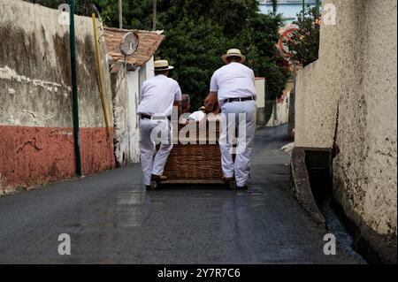 Rodelfahrer in traditionellen weißen Uniformen führen einen Korbschlitten durch die alten Straßen madeiras Stockfoto