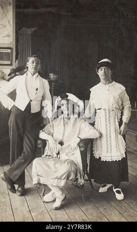 Vintage-Foto von drei Soldaten, zwei im Drag, posieren im Kostüm. Fotografische Postkarte, ca. 1920. In der Mitte sitzt ein Soldat in Drag-gekleidet als Dame, mit (links) einem Mann im weißen Smoking und (rechts) einem Mann im Drag-gekleidet als Dienstmädchen. Im Hintergrund gibt es einen Blick auf andere. Stockfoto