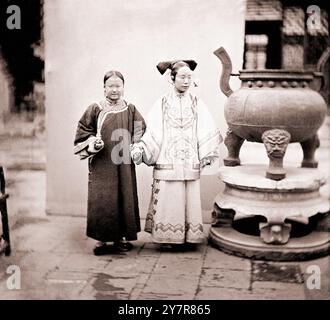 Vintage-Foto einer Mandschu-Frau mit ihrem Dienstmädchen neben einem Bronzebranke, Peking. Nord-Zhili (Pei-chih-li oder Provinz Peking), China. Von John Thomson, 1869. Zwei Frauen stehen neben einem Weihrauchbrenner, eine hält den Arm der anderen. Die Dame trägt einen Yunjian (Wolkenkragen). Während das Dienstmädchen direkt in die Kamera starrt, sieht ihre Geliebte unsicher aus und deutet darauf hin, dass Thomson versuchte, sich von der formalen Porträttradition zu entfernen und versuchte, seine Motive als Individuen darzustellen. Es zeigt auch, dass er versuchte, die mehrdeutige Rolle einer Mandschu-Magd zu vermitteln: Sie war eher wie eine Gefährtin t Stockfoto