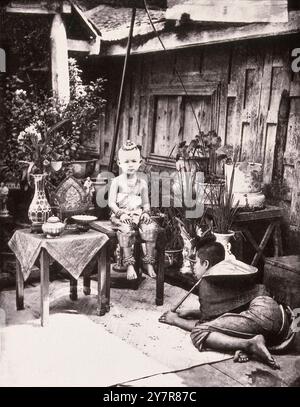 Vintage-Foto des siamesischen Prinzen. Siam (Thailand). 1865 (von John Thomson) Ein kleines Kind (weniger als fünf Jahre alt), das einen Knoten trägt, schwere Armreifen an den Handgelenken und Knöcheln, auf einem Tisch sitzt. Ein Diener (?) Er hält einen Fliegenbesen, der vor ihm auf dem Boden hockt. Stockfoto