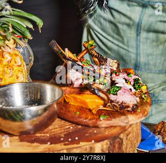 Perfekt gegrillte Lammkoteletts auf einem Holzbrett, begleitet von frischem Gemüse und garniert mit Kräutern, servierfertig. Stockfoto