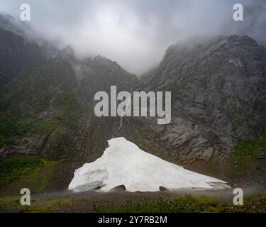 Eishöhle am Fuße eines nebeligen Berges mit einem Wasserfall, der durch felsige Klippen fließt, umgeben von Nebel und Wolken in einer abgelegenen Wildnis. Stockfoto