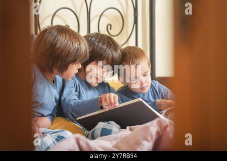 Blick durch eine halboffene Tür von drei Brüdern, die auf einem Bett in einem Schlafsaal sitzen und ein großes Buch lesen. Der ältere Bruder erzählt ihnen die Geschichte und dreht sich um Stockfoto