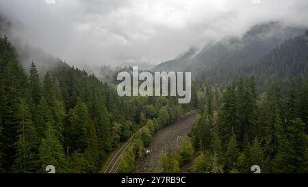 Blick aus der Vogelperspektive auf eine gewundene Straße durch einen nebeligen Wald mit einem Flussbett, umgeben von Bergen und dichten Bäumen in abgelegener Wildnis. Stockfoto