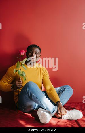 Glücklicher junger afrikanischer Mann, der eine Rose hält, während er auf dem Boden vor der roten Wand sitzt Stockfoto