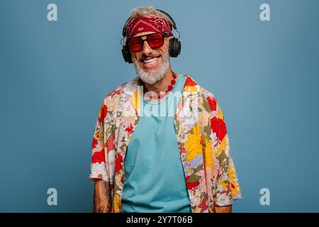 Fröhlicher Seniorenmann in funky Hemd und Kopfhörern, der die Musik vor blauem Hintergrund hört Stockfoto