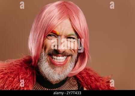 Glücklicher reifer schwuler Mann mit regenbogenfarbenen Augenbrauen mit rosa Perücke auf braunem Hintergrund Stockfoto