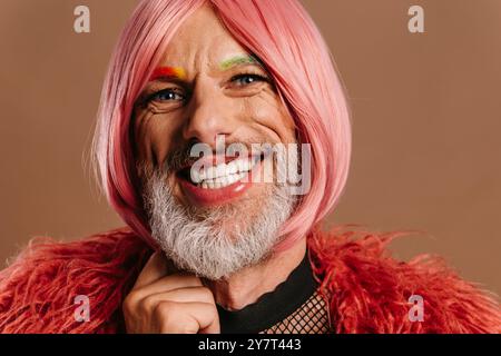 Porträt eines reiferen schwulen Mannes mit regenbogenfarbenen Augenbrauen mit rosa Perücke auf braunem Hintergrund Stockfoto