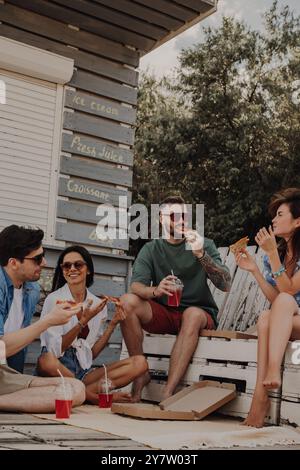 Gruppe fröhlicher Freunde, die Pizza und Getränke genießen, während sie sich in der Nähe des Strandcafés entspannen Stockfoto