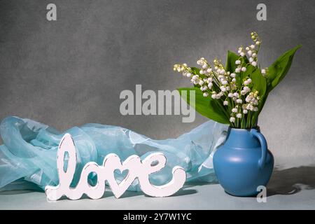 Kleiner Strauß weißer Lilien aus dem Tal auf dünnen grünen Stielen. Lily of Valley Glocken hängen von blauer Vase. Neben der Aufschrift „Love“ und dem blauen Schal. St Stockfoto