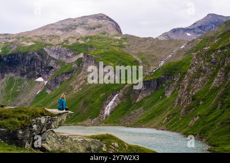 Frau sitzt auf einem großen Felsen, Blick über den blauen Bergsee, umgeben von Bergen, die von grüner Vegetation bedeckt sind. Kaukasische Frau, die nachdenklich den Kampf genießt Stockfoto