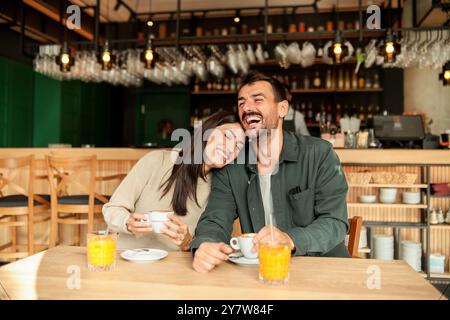 Ein Paar teilt sich einen freudigen Moment in einem stilvollen Café, schlürft Kaffee und genießt hellen Orangensaft, während es gemeinsam lacht, umgeben von einem einladenden Restaurant Stockfoto