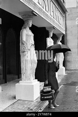 Mit einem Regenschirm über dem Kopf hat diese junge Dame nichts im Kopf, aber nicht die Statuen hinter sich Wie ein griechischer Tempel, verwendet dieser neue Antiquitätenladen in Kensington sechs exakte Nachbildungen der „Caryatiden“, um seine Fassade zu stützen. Die ursprünglichen „Karyatiden“ wurden verwendet, um das Dach eines der Tempel auf der Akropolis in Athen zu stützen. Dieser neue Laden mit seinen Ladenläden soll in etwa zwei Wochen eröffnen. Antiquitätenmarkt, Kensington High Street, London, England. 14. September 1967 Stockfoto