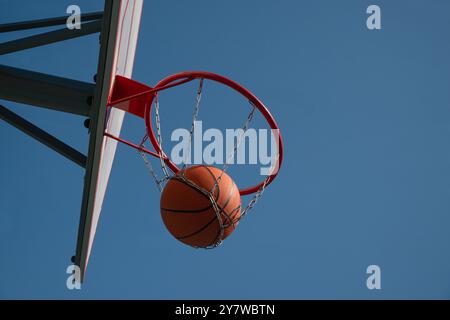 Orangener Basketballball im Basketballkorb vor klarem blauem Himmel. Spielmoment. Im Freien. Kopierbereich. Stockfoto