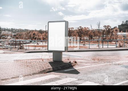 Eine leere, vertikale Werbetafel mit einem leeren weißen Display steht an einer Straßenecke. Der Hintergrund zeigt eine sonnige urbane Umgebung mit Autom Stockfoto