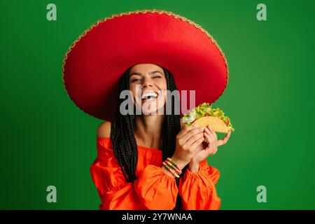 Glückliche junge mexikanische Frau in Sombrero, die Taco hält und vor grünem Hintergrund lächelt Stockfoto