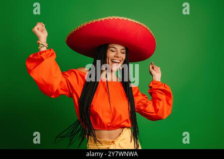 Schöne junge mexikanische Frau in Sombrero, die glücklich aussieht, während sie vor grünem Hintergrund tanzt Stockfoto