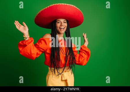 Attraktive junge mexikanische Frau in Sombrero, die glücklich aussieht, während sie vor grünem Hintergrund tanzt Stockfoto
