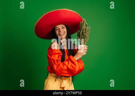Schöne junge mexikanische Frau in Sombrero, die Kakteen hält und vor grünem Hintergrund lächelt Stockfoto