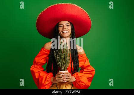 Fröhliche junge mexikanische Frau in Sombrero, die Kakteen vor grünem Hintergrund hält Stockfoto