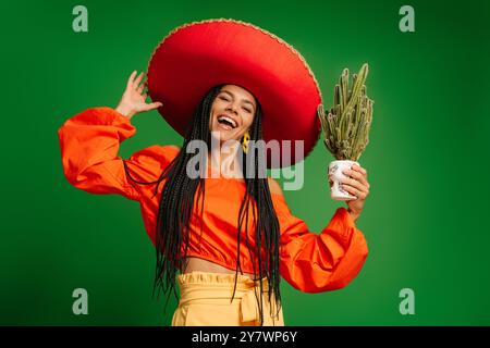 Attraktive junge mexikanische Frau in Sombrero, die Kakteen hält und vor grünem Hintergrund lächelt Stockfoto