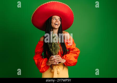 Fröhliche junge mexikanische Frau in Sombrero, die Kakteen vor grünem Hintergrund hält Stockfoto