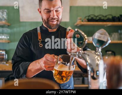 Ein lächelnder, stilvoller Bärenbarmann in schwarzer Uniform mit einer Schürze, der frisches Lagerbier in einen Glasbecher an der Bar zapft. Erfolgreiche Leute, Bierkonzern Stockfoto