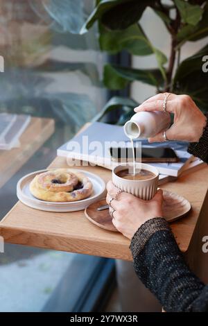 Weibliche Hände geben Milch zu einem Cappuccino Latte auf einem schönen Coffee Shop Tisch. Seitenansicht Stockfoto