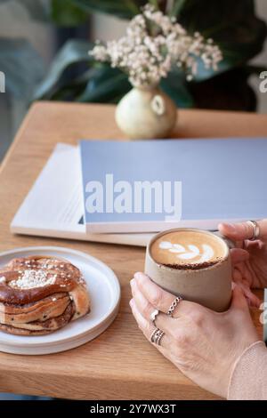 Die Frau hält Kaffee mit den Händen auf einem Tisch in einem Café, umgeben von köstlichem Gebäck und Zeitschriften. Stockfoto