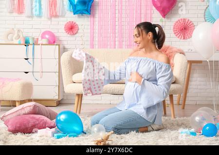 Schöne glückliche schwangere Frau mit Babykleidung zu Hause. Gender Reveal Party Konzept Stockfoto