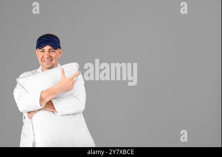 Schöner reifer glücklicher Mann im Pyjama und Bademantel mit Kissen, der auf etwas auf grauem Hintergrund zeigt Stockfoto