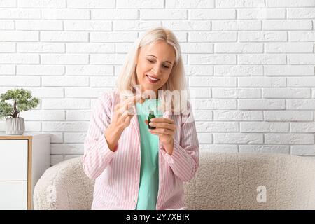 Reife Frau mit einer Flasche ätherischen Öls, die auf dem Sofa in der Nähe einer hellen Ziegelwand sitzt Stockfoto