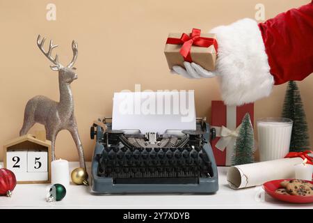 Weihnachtsmann-Hand mit Weihnachtsdekor, Geschenkboxen, Glas Milch und Schreibmaschine am Holztisch Stockfoto